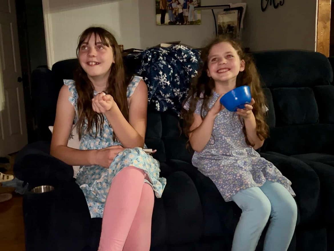 two sisters eat popcorn and watch a movie on the couch
