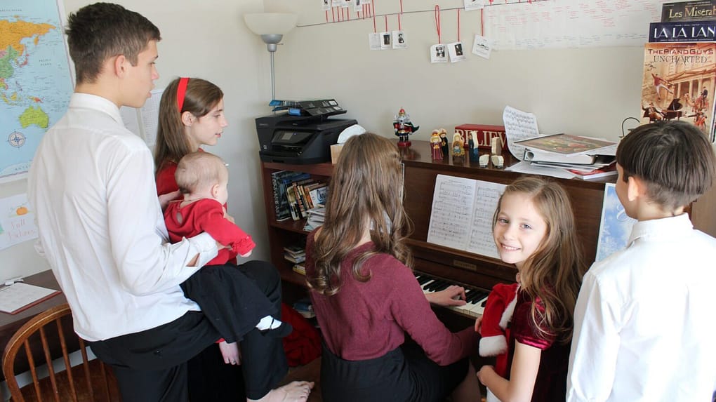 Five siblings gather around the piano to sing Christmas carols.