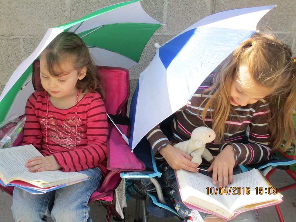 Two little girls read books outside in chairs and holding umbrellas