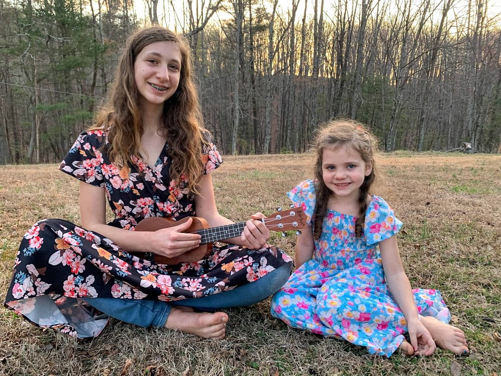 two sisters sitting on the grass in dresses playing the ukulele