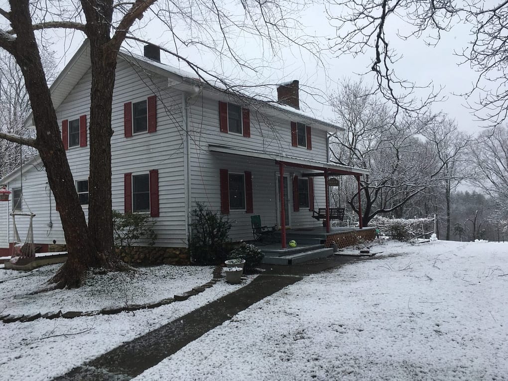 White farmhouse with red shutters in a snowy landscape scene.