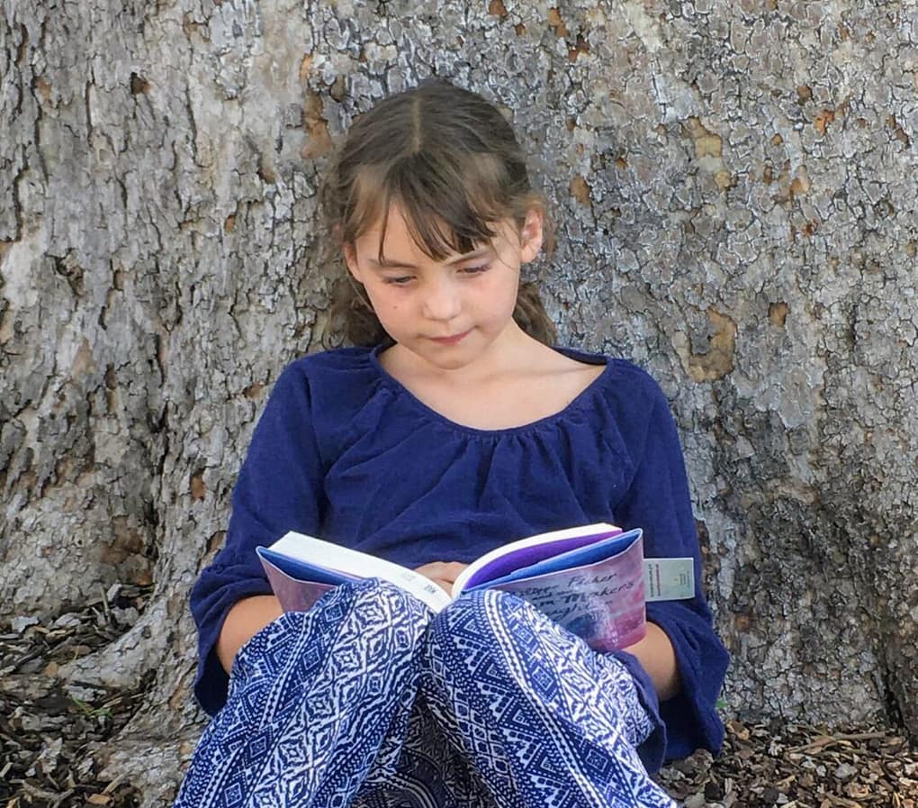 girl in a blue shirt reading a book outside leaning against a tree