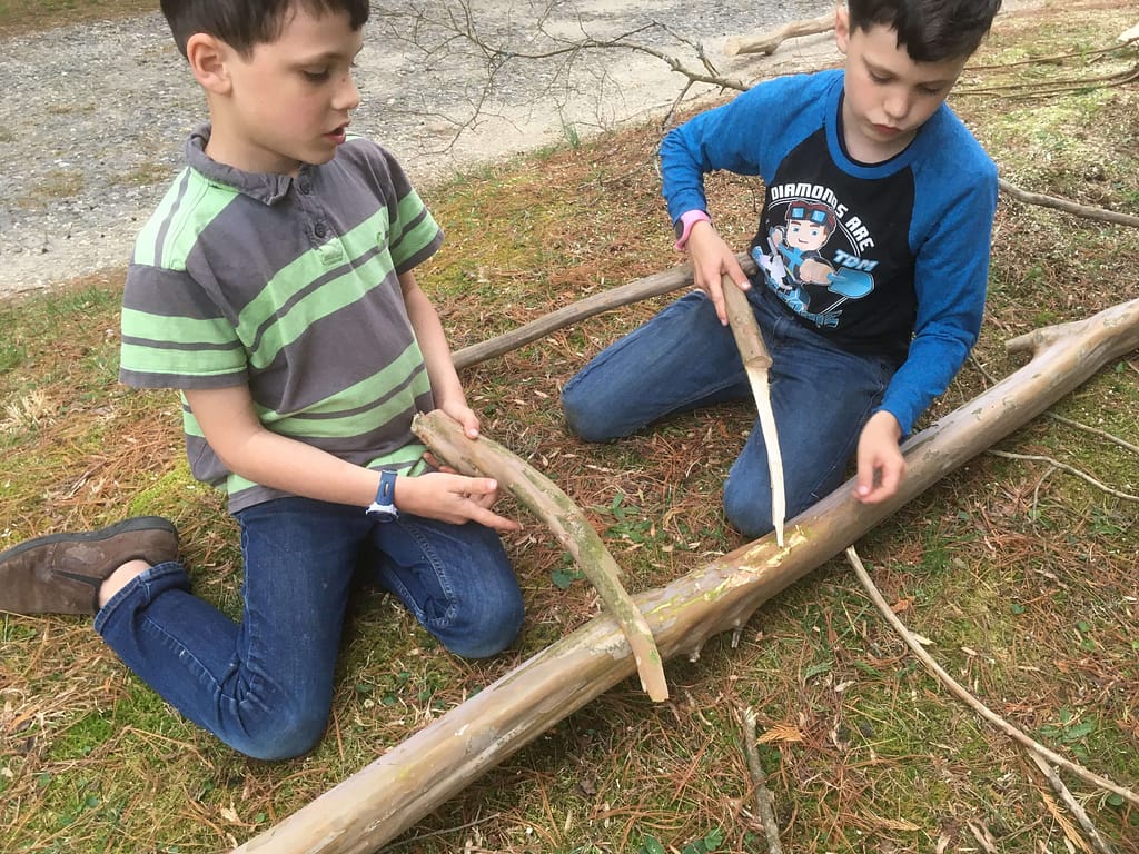 Two boys using sticks for pretend saws to saw a branch on the ground