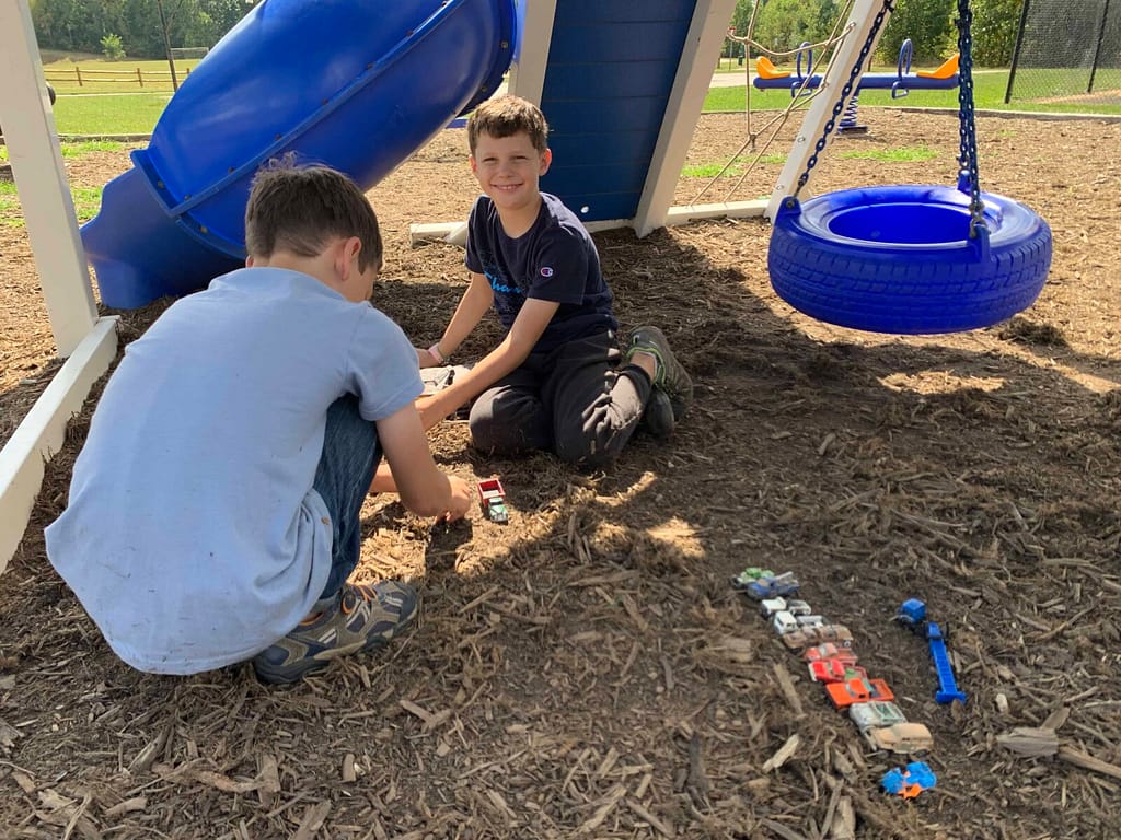 two boys playing cars in the dirt at the park