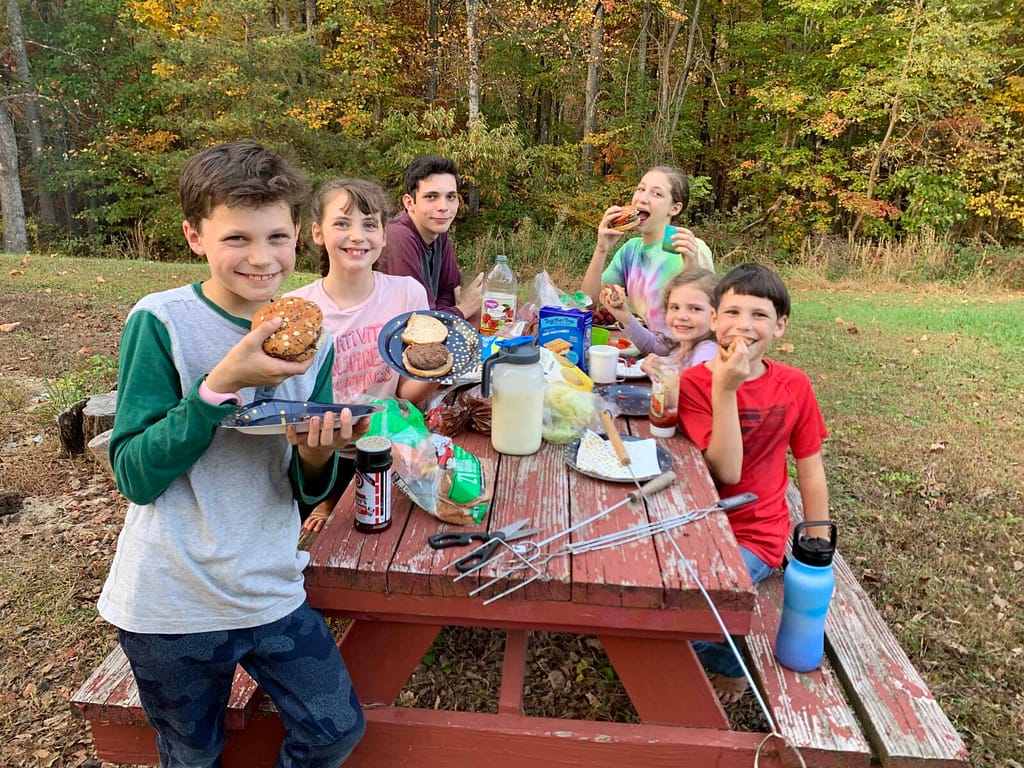 family of 6 children eating hamburgers and hotdogs at picnic table