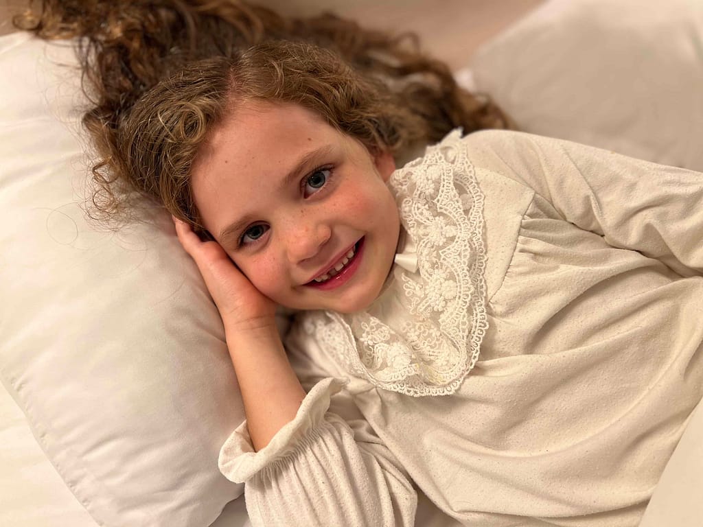 7 year old girl in white nightgown lying in bed and smiling