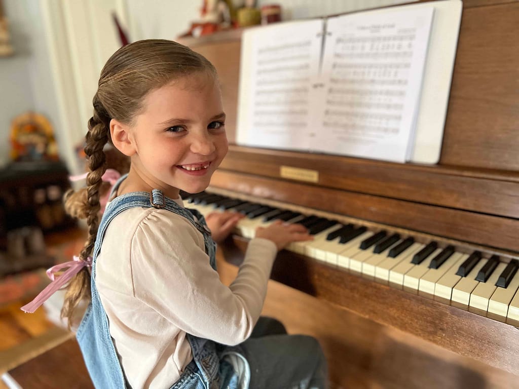 girl in overalls and braids smiling while playing the piano