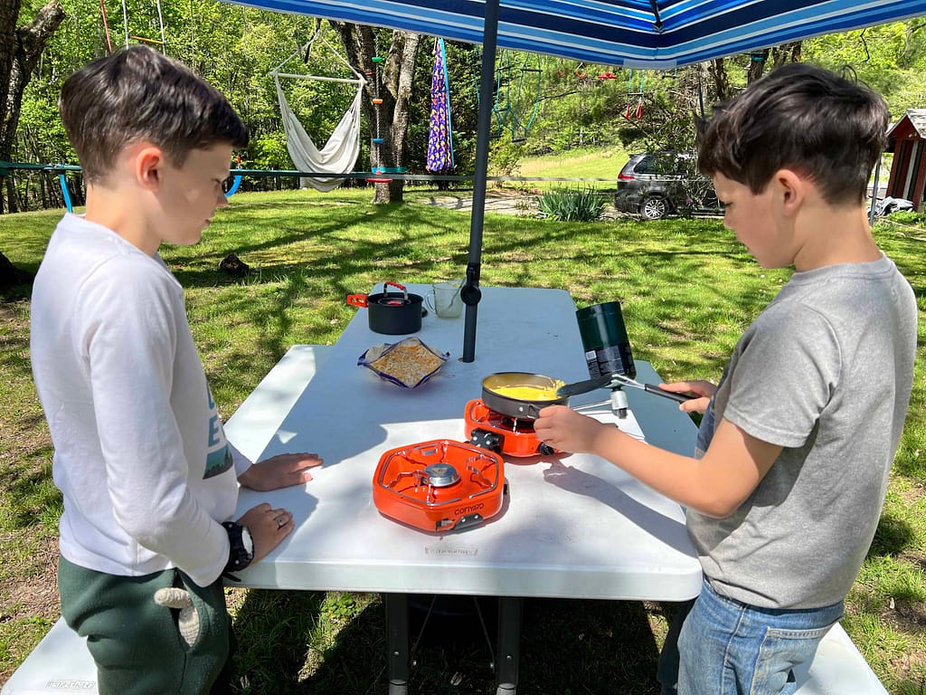 2 boys cook eggs over camp stove