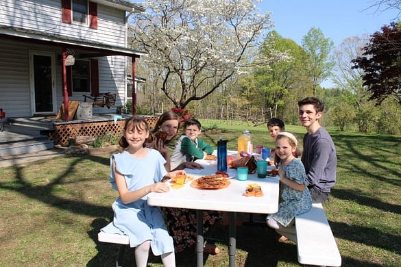 family of 6 kids eat cheese sandwiches at a picnic table in their front yard.