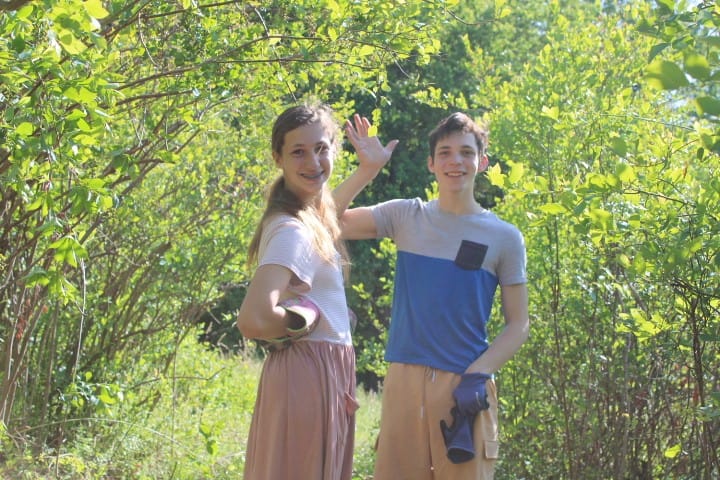 two teenage siblings wave at the camera while they're working outside