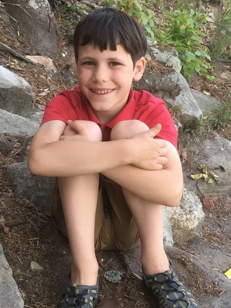 Boy in shorts and t-shirt sitting in the dirt and smiling at Pilot Mountain