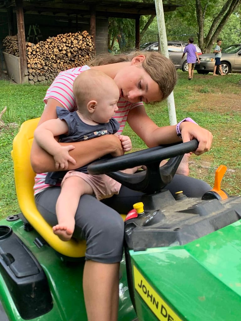13 year old on a riding mower holding a baby